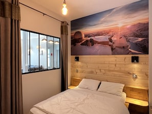 Un dormitorio con una cama grande, una ventana con cortinas, cabecera de madera y una lámpara fijada a la pared.