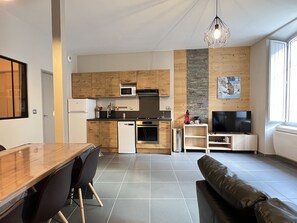 A modern kitchen with wooden cabinets, a microwave, and a dishwasher. There is a dining area with a table and chairs, and a living room with a sofa and a television. - Joli T3 rénové ds une villa 1900 (Cauterets)