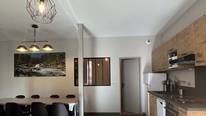 A modern kitchen with a dining area, featuring a large table and chairs, a hanging light fixture, and a view of a forest stream.