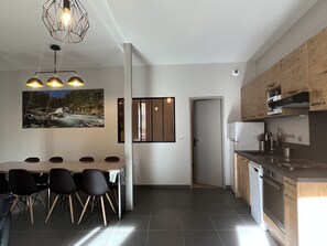 A modern kitchen with a dining area, featuring a large table and chairs, a hanging light fixture, and a view of a forest stream.