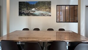 A conference room with a wooden table and black chairs, a painting of a river, and pendant lighting.