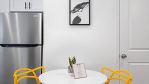 A compact kitchen with a stainless steel refrigerator, a white table with two yellow chairs, and a framed picture on the wall.