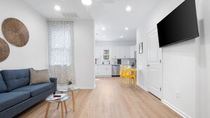A modern living room with a sofa, coffee table, and yellow chairs. A kitchen is visible in the background with white cabinets and a black TV mounted on the wall.