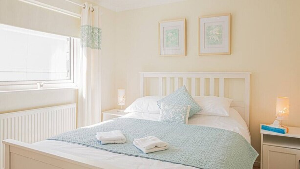 Une chambre à coucher bien rangée, avec un lit, deux tables de chevet, une fenêtre et des tableaux encadrés accrochés au mur.