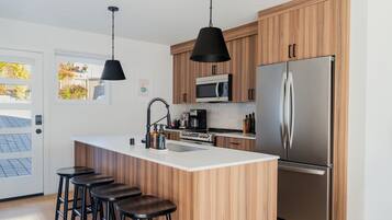 A modern kitchen with wooden cabinets, a stainless steel refrigerator, a microwave, and a bar area with stools.