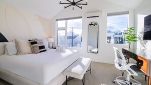 Une chambre moderne avec un grand lit, un bureau et une chaise. Il y a une fenêtre donnant sur des immeubles, un miroir et une plante.