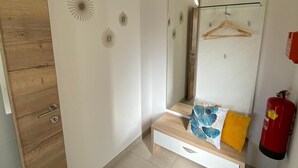 A bedroom with a wooden wardrobe, a mirror, a bedside table with a blue and white pillow, a red fire extinguisher, and a blue furry rug.