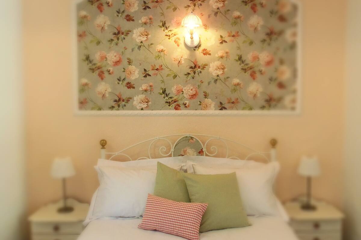 A bedroom with a floral wallpaper, a bed with white and green pillows, and two bedside tables with lamps.
