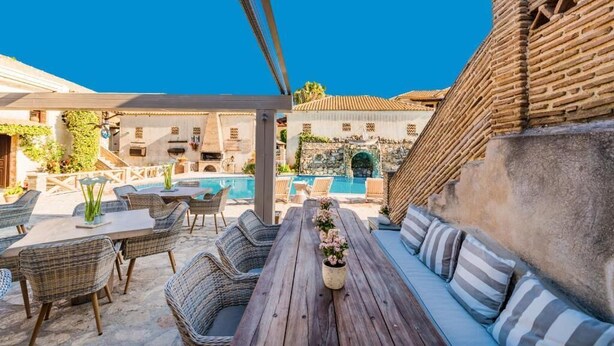 Un espace extérieur aménagé avec des meubles en osier, une table en bois ornée d’un vase de fleurs, et une vue sur une piscine et des bâtiments en pierre.
