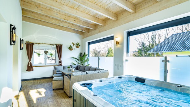 A modern indoor swimming pool area with a wooden ceiling, a hot tub, and a seating area with a sofa and table.