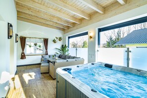 A modern indoor swimming pool area with a wooden ceiling, a hot tub, and a seating area with a sofa and table.