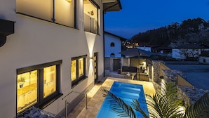 A modern two-story house with a swimming pool, outdoor seating, and a view of the surrounding area.