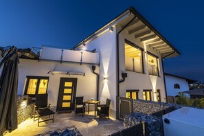 A modern two-story house with a patio area featuring a table and chairs, a stone wall, and a covered outdoor space.