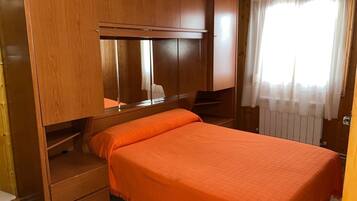 Un dormitorio pequeño con una cama individual, armario de madera y ventana con cortinas traslúcidas.