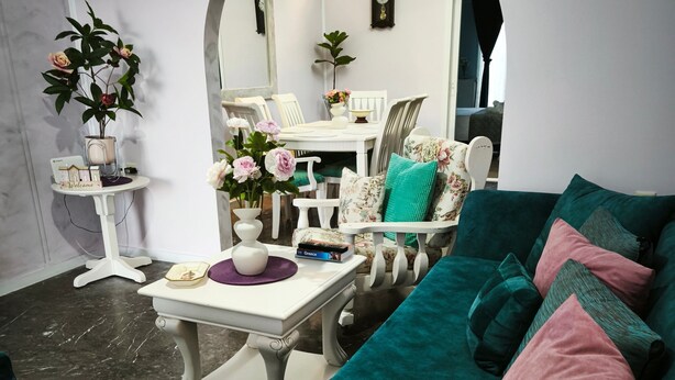 A living room with a teal sofa, a white coffee table, and a floral armchair. There is a white side table with a vase of flowers and a clock on the wall. - Vacation Home "Marilidia House" with 2 Balconies, Shared Terrace & Wi-Fi (Kastelli)