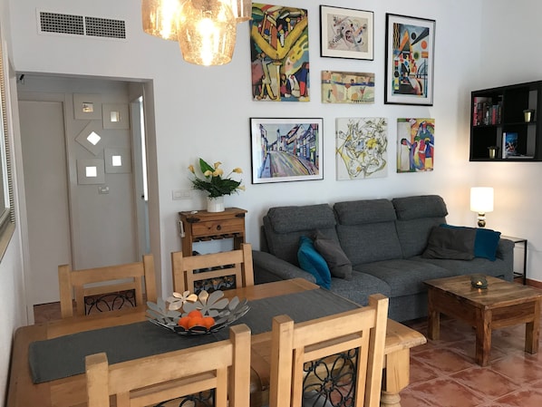 Una sala de estar con un sofá gris, una mesa de comedor de madera, sillas, una pequeña mesita auxiliar de madera y una pared decorada con diversas obras de arte enmarcadas.