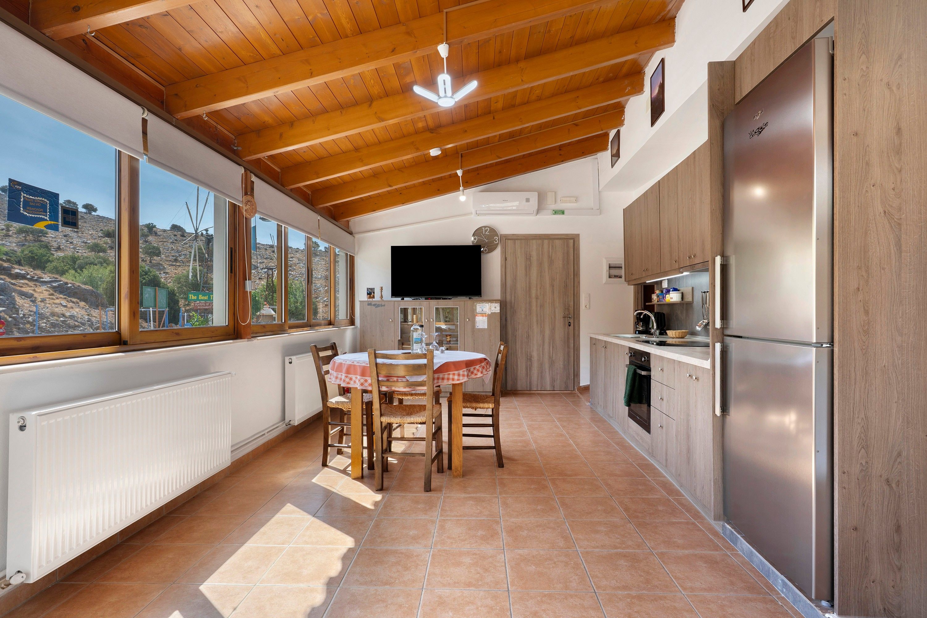 A kitchen with wooden ceiling beams, a dining table with chairs, a built-in oven, and a large window overlooking a rocky landscape.