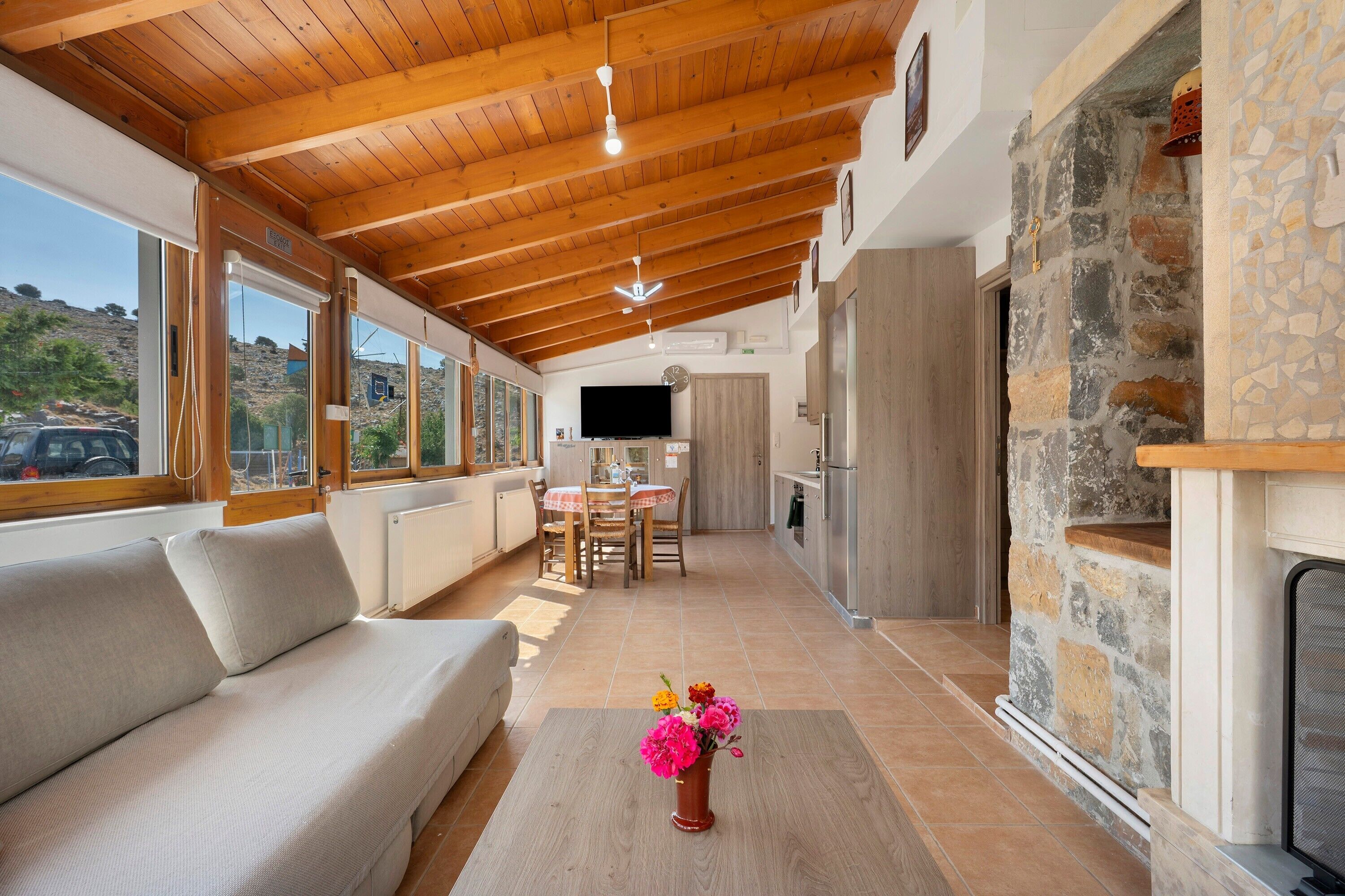 A spacious living area with a wooden ceiling, a sofa, a dining table with chairs, and a stone wall.