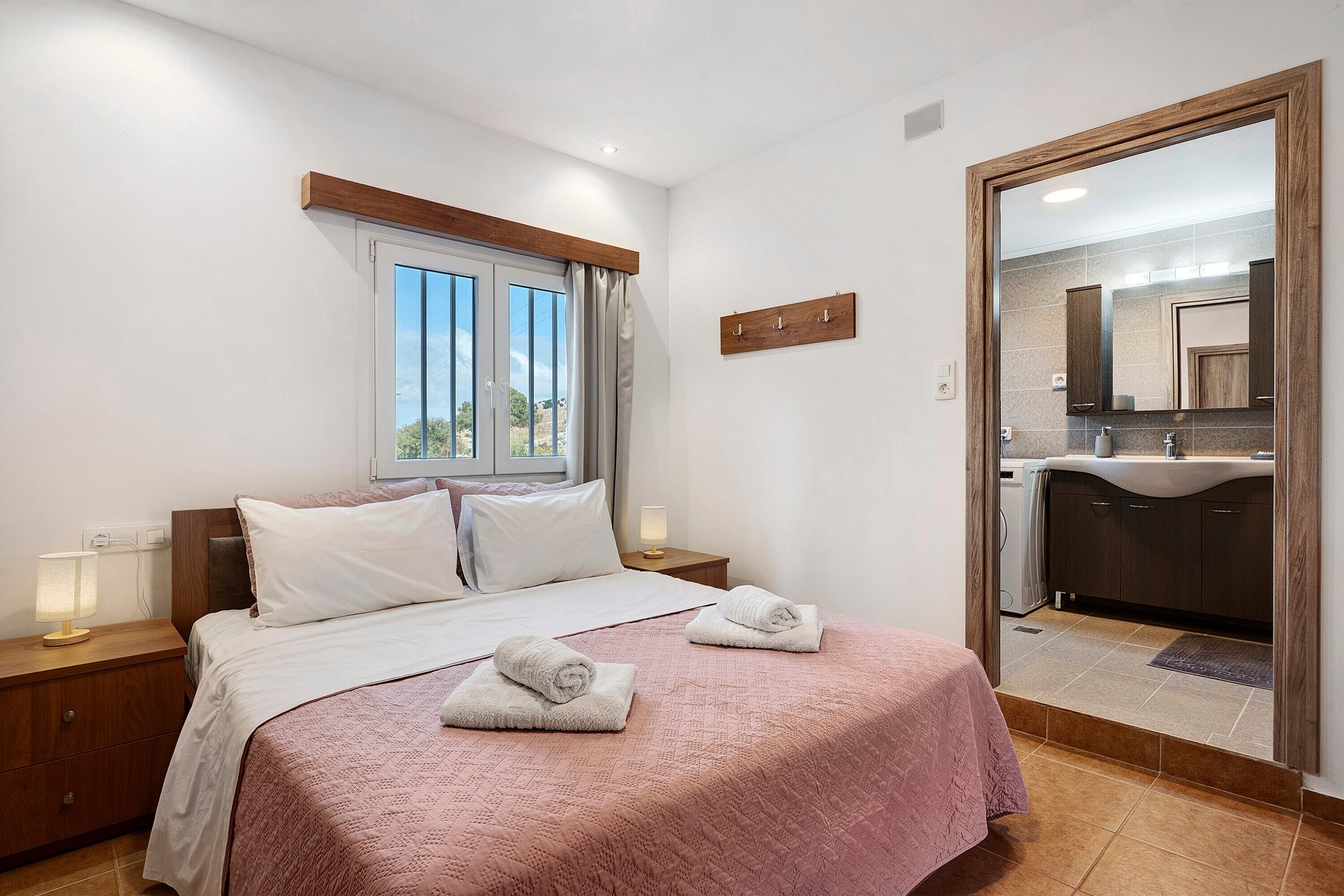 Une chambre à coucher avec un lit, des tables de chevet, une fenêtre et une salle de bain visible à travers une porte ouverte.