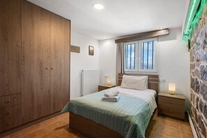 Ein Schlafzimmer mit großem Kleiderschrank, einem Bett mit grüner Tagesdecke, zwei Nachttischen inklusive Lampen, einem Fenster mit Vorhängen und einem Heizkörper.