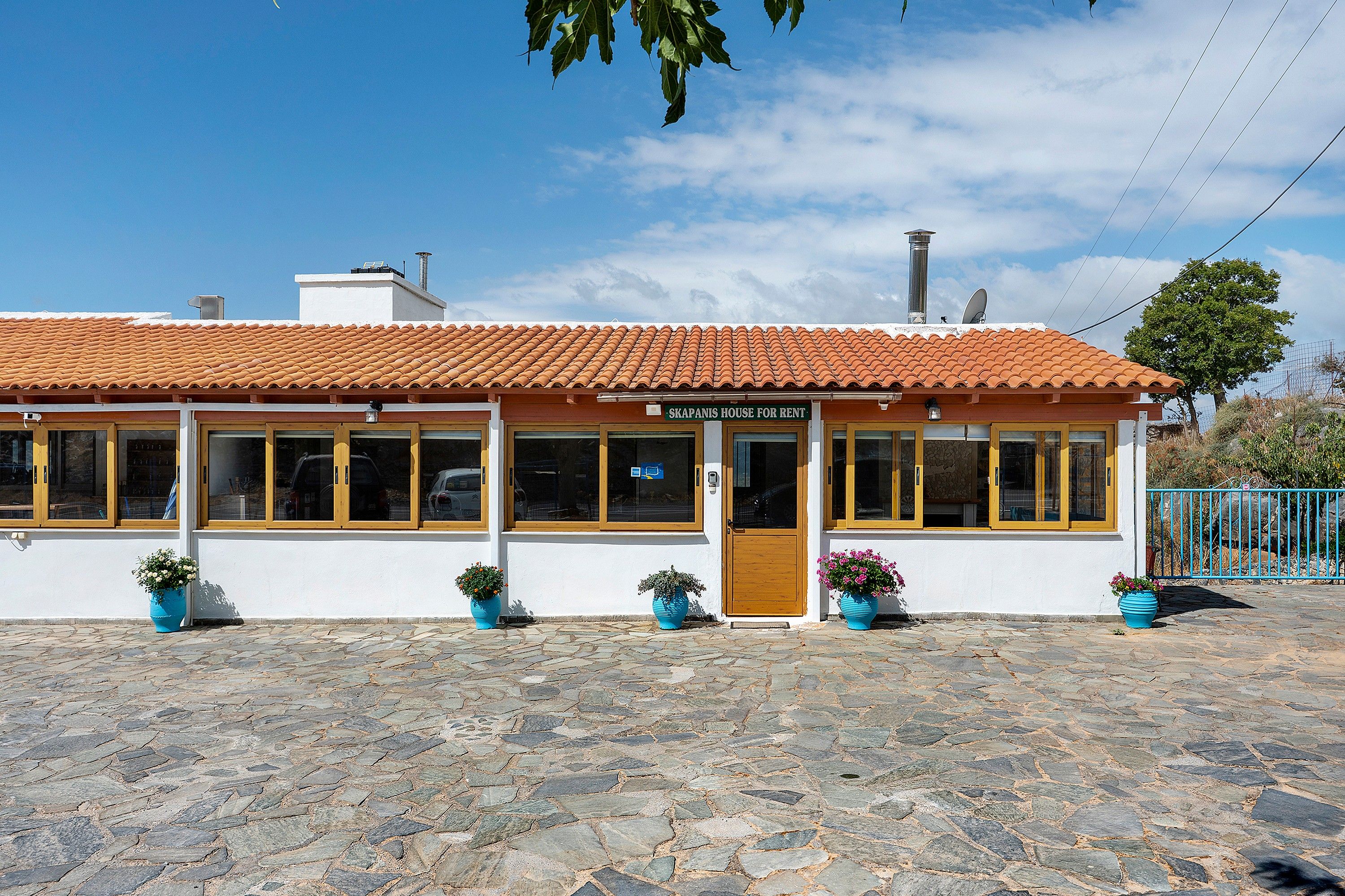 A two-story building with a tiled roof, large windows, and a sign that reads 'SKAPANIS HOUSE FOR RENT'.