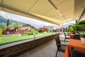 Une terrasse extérieure avec un sol en pierre, des meubles en osier et une vue sur un jardin aménagé et des montagnes lointaines.