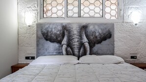 Ein Bett mit weißer Bettdecke, darüber ein großes Elefantenbild an der Wand und ein Fenster mit geometrischem Muster.