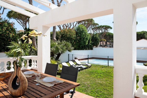 An outdoor patio with a table set for dining, a white fence, and a pool in the background.