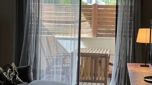 A room with a sofa, a small table, a lamp, and a view through a glass door to an outdoor area with a wooden deck and chairs.
