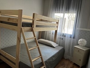 A bunk bed with a ladder, a bedside table, and a window with sheer curtains.