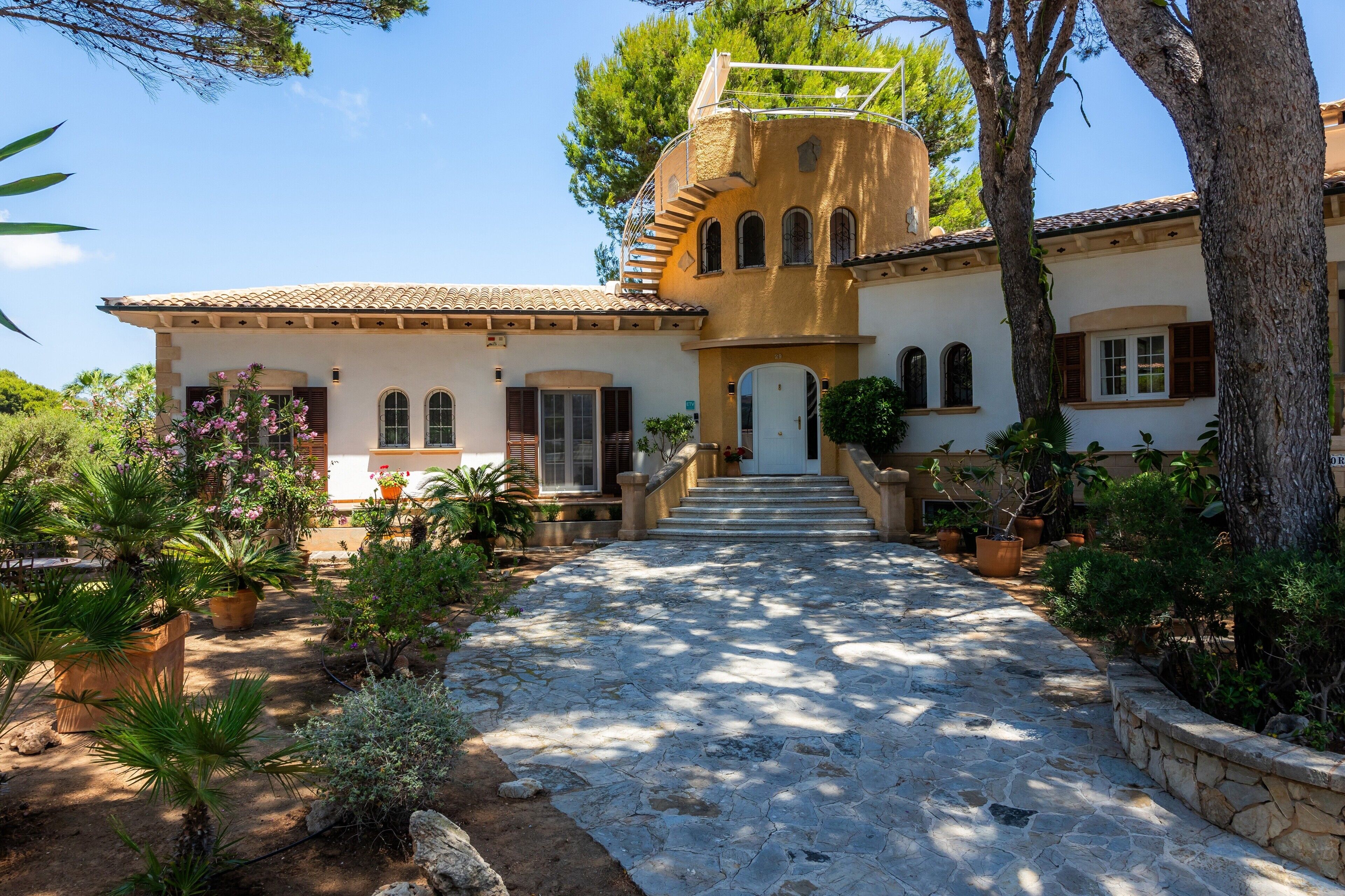 Un bâtiment de style méditerranéen, avec un chemin de pierre menant à l’entrée, entouré de plantes en pot et d’arbres.