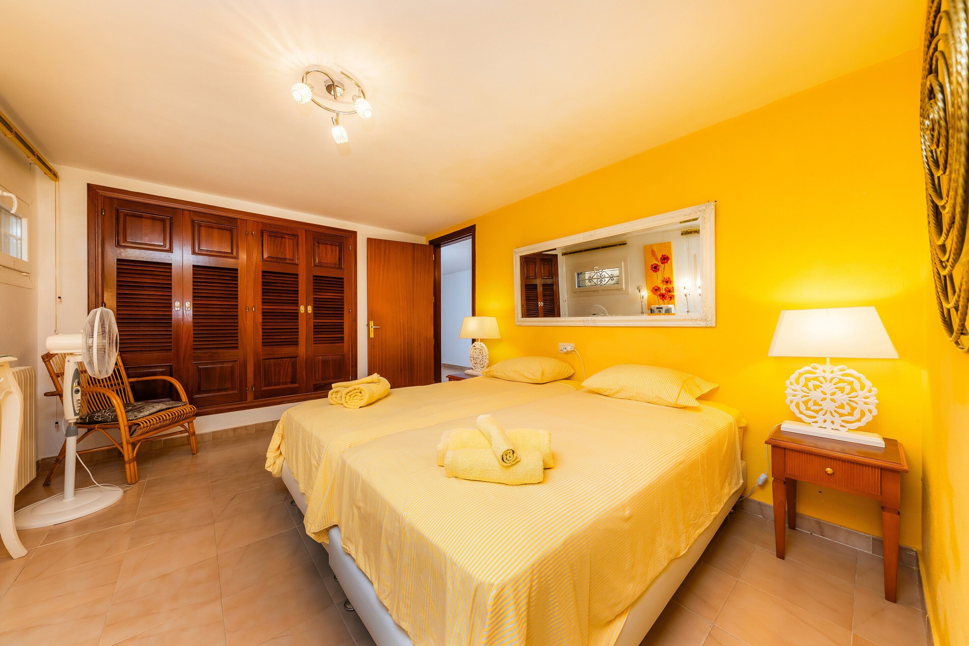 A bedroom with two beds, a wooden wardrobe, a chair, a bedside table, and a mirror.