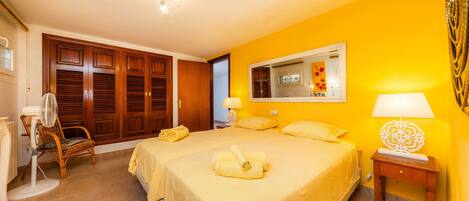 A bedroom with two beds, a wooden wardrobe, a chair, a bedside table, and a mirror.