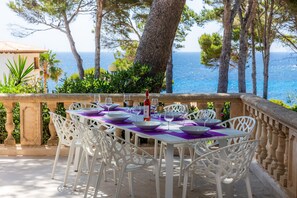 Un área de comedor al aire libre con sillas y mesas blancas, un balcón de piedra y vista al mar.