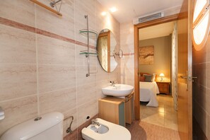 A modern bathroom with a toilet, sink, and mirror. There is a wooden cabinet and a bedside table with a lamp.