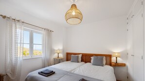 Une chambre à coucher avec un lit, deux tables de chevet avec des lampes, une fenêtre avec des rideaux et un luminaire suspendu.