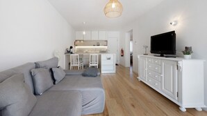 Un salon moderne avec un canapé gris, un meuble de rangement blanc pour les appareils électroniques et, au fond, un espace cuisine.