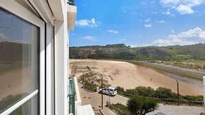 Une vue sur la plage depuis un balcon, avec une voiture blanche garée sur le sable, un ciel dégagé et un paysage vallonné en arrière-plan.