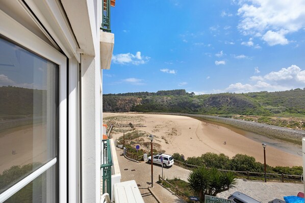 A beach view from a balcony with a white car parked on the sand, a clear sky, and a hilly landscape in the background.
