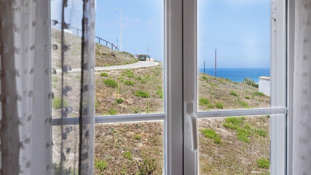 Une vue sur la côte, aperçue à travers une fenêtre ornée d’une rambarde en métal, avec un sentier qui mène à la mer.