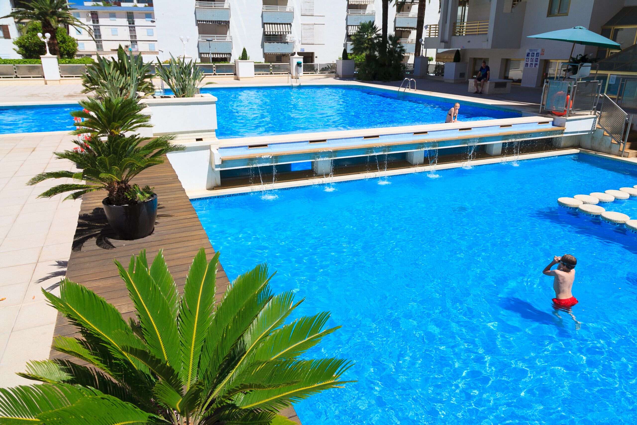 A swimming pool with a wooden deck, surrounded by potted plants and a building in the background.