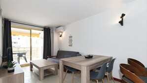 A modern living room with a wooden dining table, grey sofa, and a small kitchen area with a plant. - UHC NOVELTY APARTMENTS 144 (Salou)