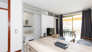 A modern hotel room with a bed, a desk with a TV, a small plant, and a view of the outside balcony. - UHC NOVELTY APARTMENTS 144 (Salou)