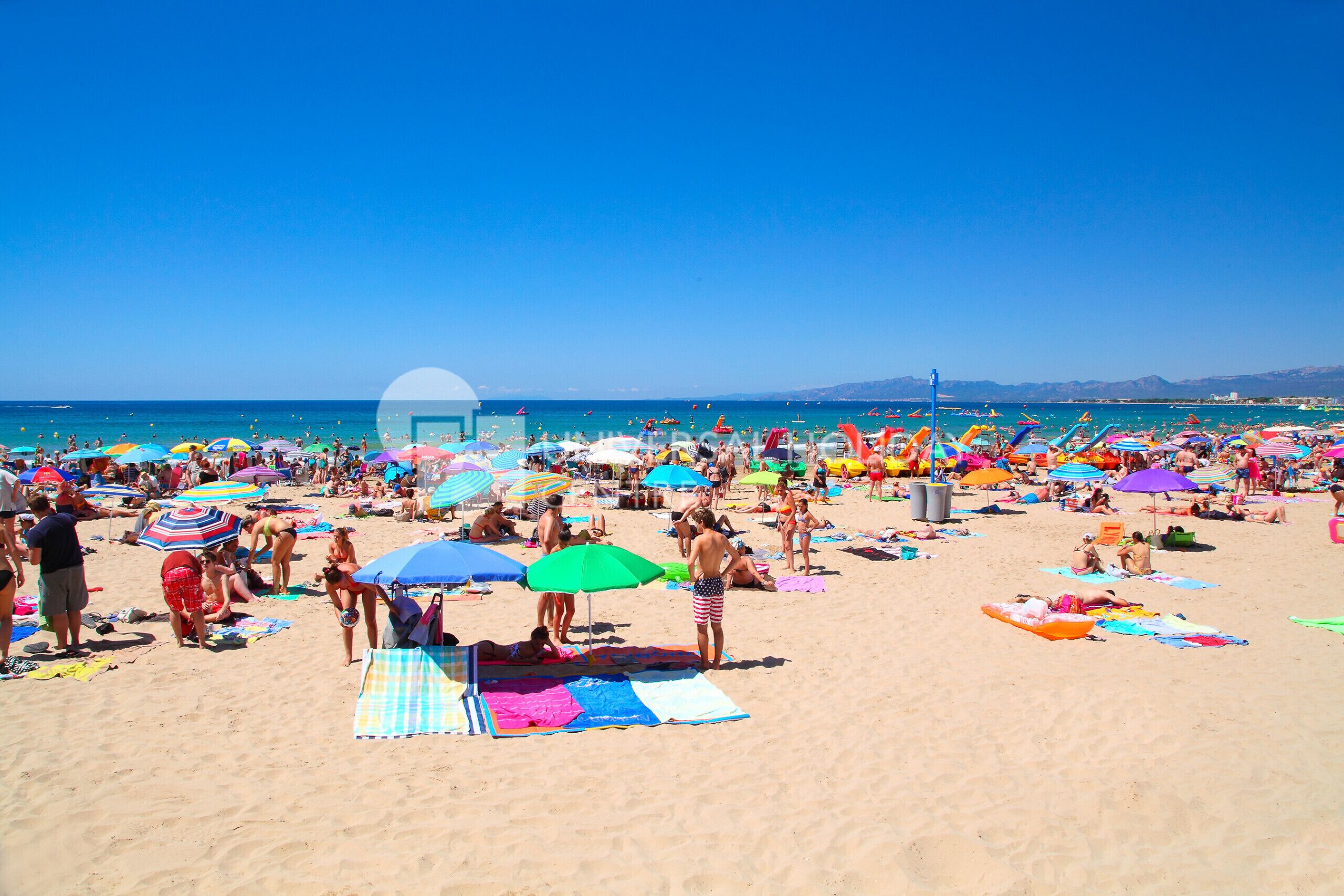 Een druk strand met talloze kleurrijke parasols en mensen die aan het zonnen zijn.