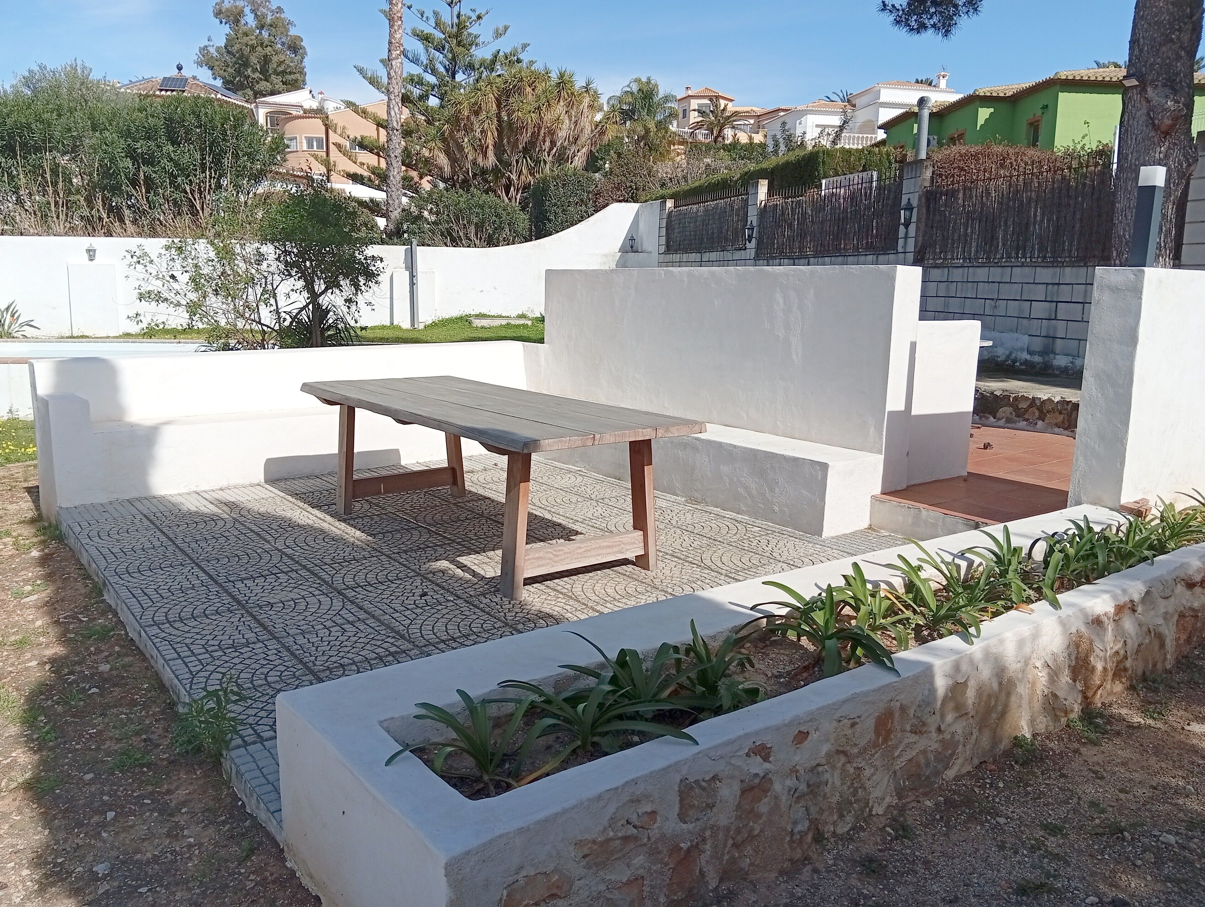An outdoor seating area with a wooden table and benches, surrounded by a white wall and a garden bed with plants.