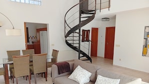 Un salon avec un canapé, une table à manger entourée de chaises, un escalier en colimaçon et, au fond, un coin cuisine.