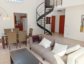 A living room with a sofa, a dining table with chairs, a spiral staircase, and a kitchen area in the background.