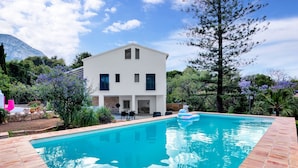 Une maison moderne à deux étages, dotée d’une piscine, entourée de verdure et avec une montagne en toile de fond.