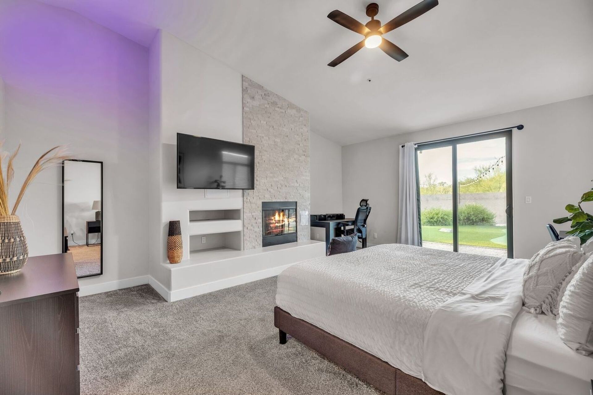 A bedroom with a fireplace, a TV, a ceiling fan, and a large window.
