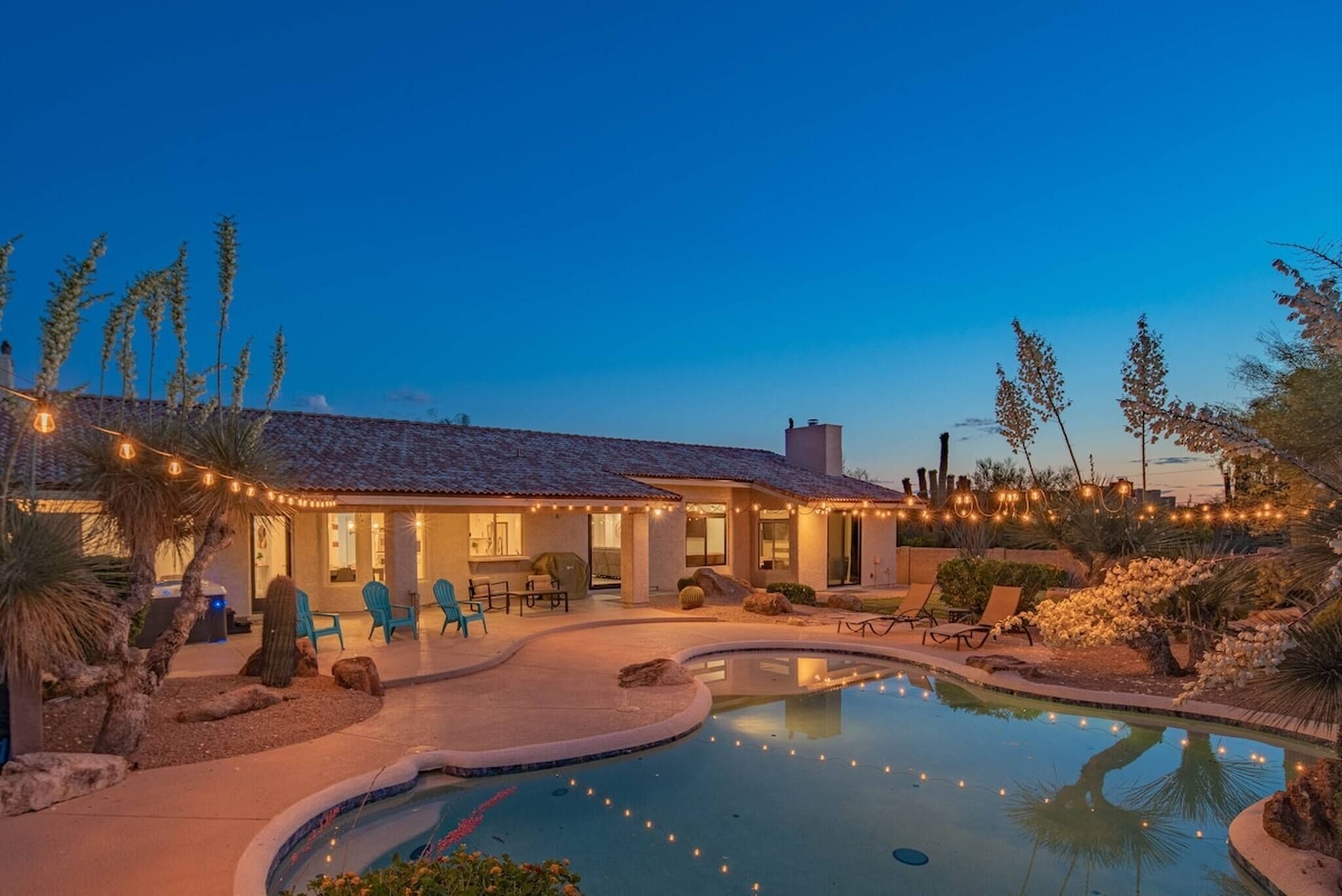 A pool with a curved edge, a patio area with chairs, and a building with string lights.